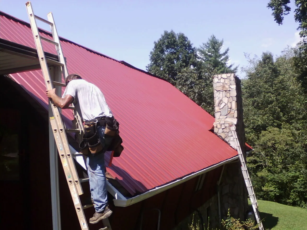 Roofer on a ladder beside a red metal roof and stone chimney — Soffit & Fascia Repair by Roofing Brothers in Demotte